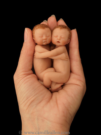 Two miniature sculpted newborn twins cuddling together in the palm of a hand